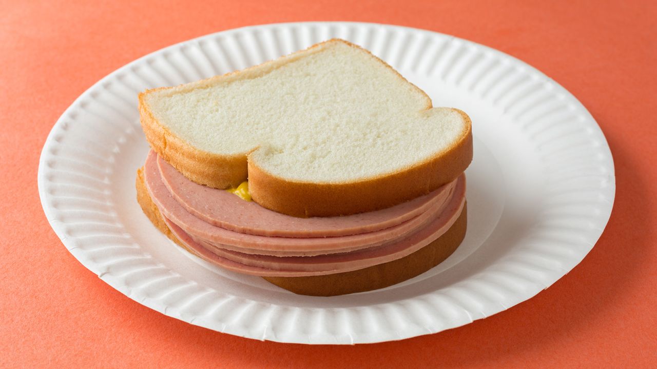 A several slices of bologna sandwich with mustard and white bread on a paper plate atop an orange table top.