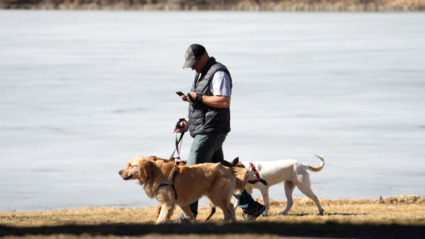 A dog walker checks a mobile device while guiding dogs to take advantage of temperatures near 60 degrees Fahrenheit while passing by an ice-covered lake in Washington Park Tuesday, Feb. 21, 2023, in Denver. Forecasters are predicting that a dangerous winter storm packing heavy snows and high winds will sweep over the intermountain West late Tuesday and Wednesday, prompting the issuing of snow advisory warnings across the region. (AP Photo/David Zalubowski)