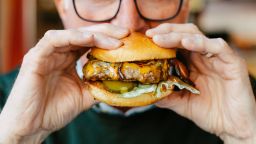 Close up image depicting a man holding a fresh burger loaded with beef and crispy bacon and salad. He is sitting in the restaurant and his eyes are wide with amazement at the size of the burger. Room for copy space.