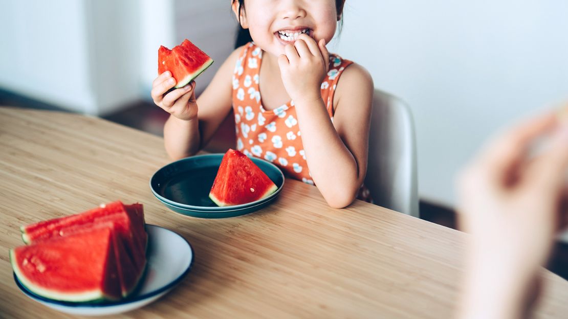 Adorable little Asian girl and mother enjoying juicy slice of fresh watermelon together sitting at the dining table at home. Enjoying summer while eating sweet juicy red fruit