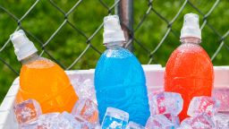 A red and white ice chest full of ice and several plastic bottles of colorful ice cold sports drinks on a hot Summer day with a chain link fence in the background.