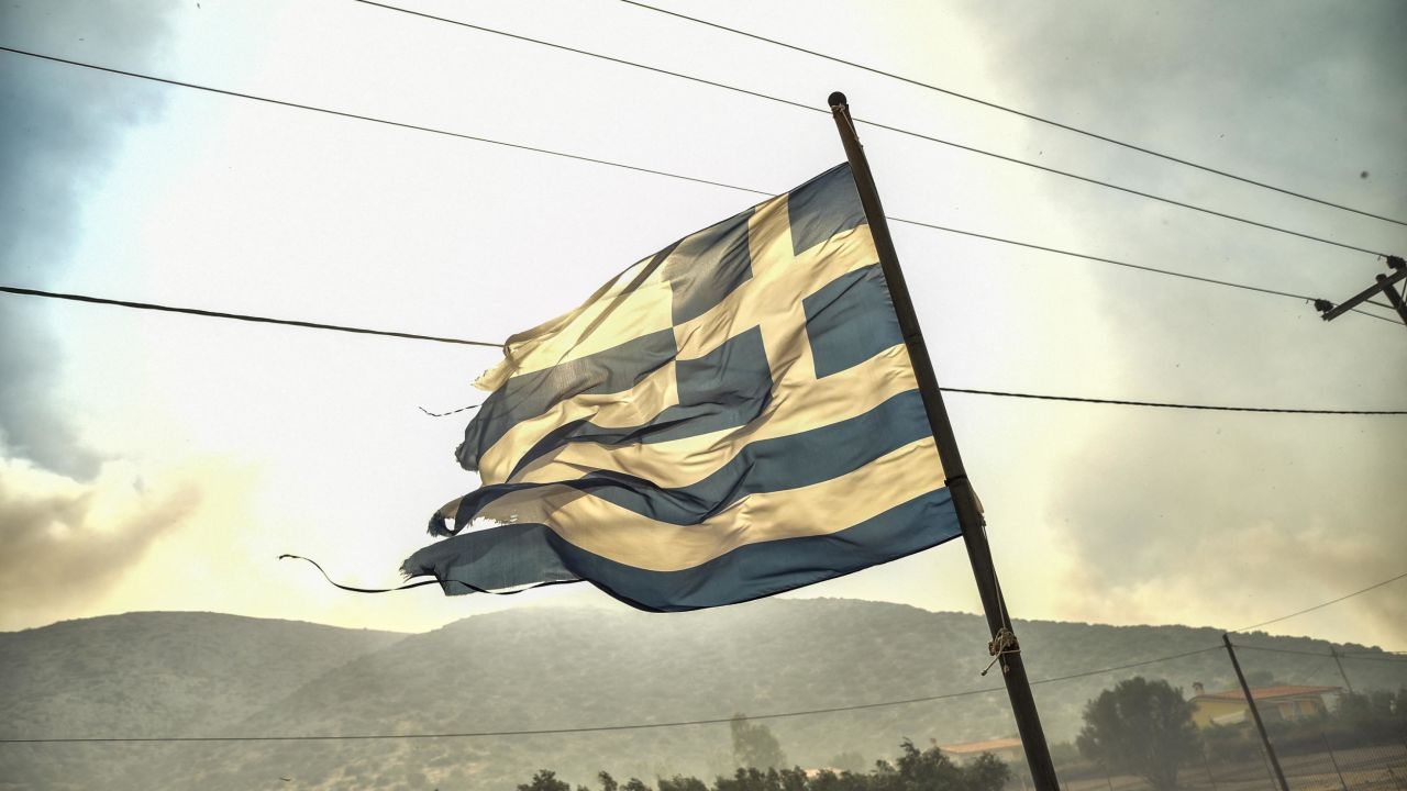 A Greek flag flutters in the wind during a wildfire some 50km southeast from the centre of Athens on July 17, 2023. A wildfire broke out near Athens on July 17 as a heatwave grips the country, firefighters said, with several seaside resorts ordered to evacuate as a precaution. The blaze started in Kouvaras, 50 kilometres (30 miles) southeast of Athens and quickly through brush and spread south toward the Attica region and the resorts of Lagonissi, Anavyssos and Saronida. (Photo by Spyros Bakalis / AFP) (Photo by SPYROS BAKALIS/AFP via Getty Images)