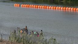 Migrants walk by a string of buoys placed on the water along the Rio Grande border with Mexico in Eagle Pass, Texas, on July 16, 2023. The buoy installation is part of an operation Texas is pursuing to secure its borders, but activists and some legislators say Governor Greg Abbott is exceeding his authority. (Photo by SUZANNE CORDEIRO / AFP) (Photo by SUZANNE CORDEIRO/AFP via Getty Images)