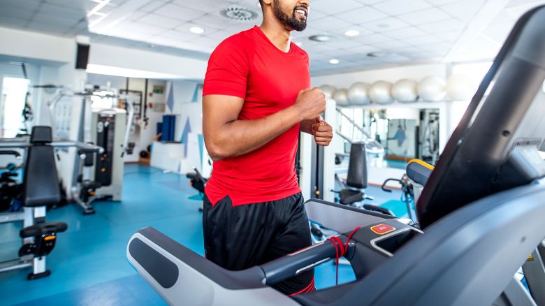 African man in early 30s wearing red t-shirt and black shorts smiling as he enjoys workout on treadmill at modern health club.