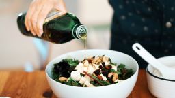 Close-up view of a bowl of fresh green salad with mozzarella, mixed nuts and dry fruits. A woman's hand was pouring olive oil into the salad.
