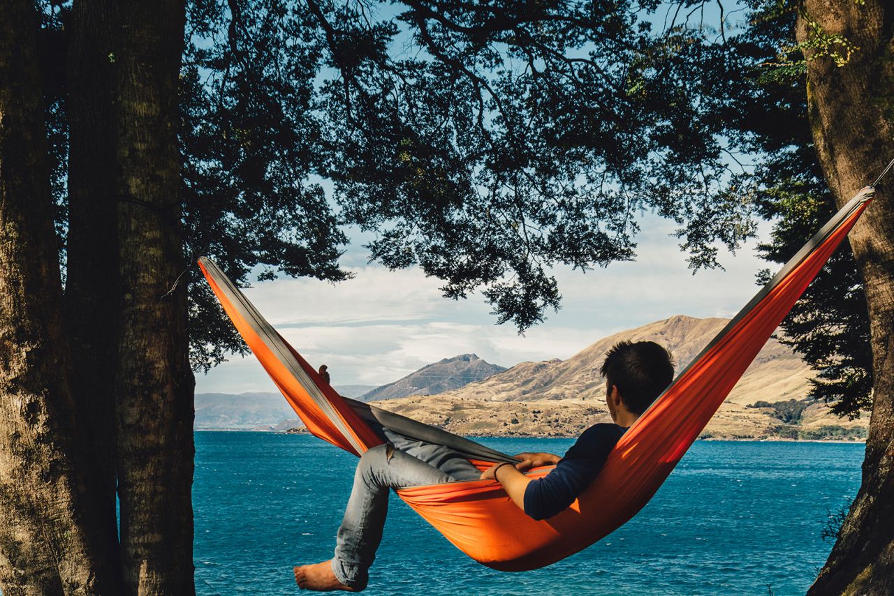 Photo shows a man relaxing in a hammock next to Lake Hawea in New Zealand.