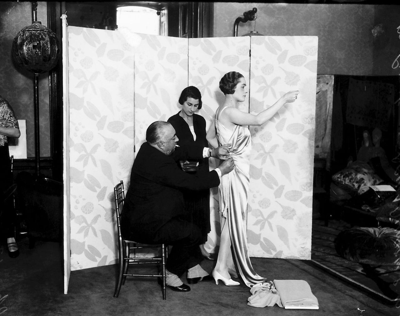 The fashion designer Paul Poiret is pictured in his studio with a fit model, as he drapes a bias-cut silk model onto her body. He is being helped by an assistant.