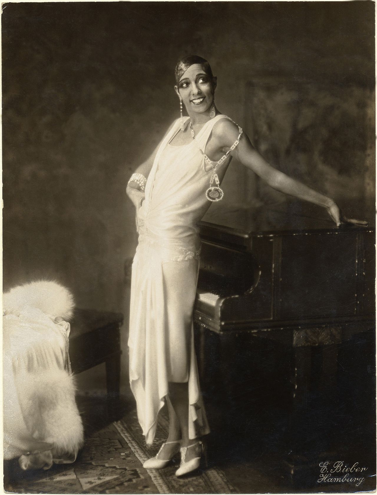 Iconic performer Josephine Baker poses in a circa-1924 promotional portrait. She is wearing a loosely-draped white flapper dress and leaning against a piano.