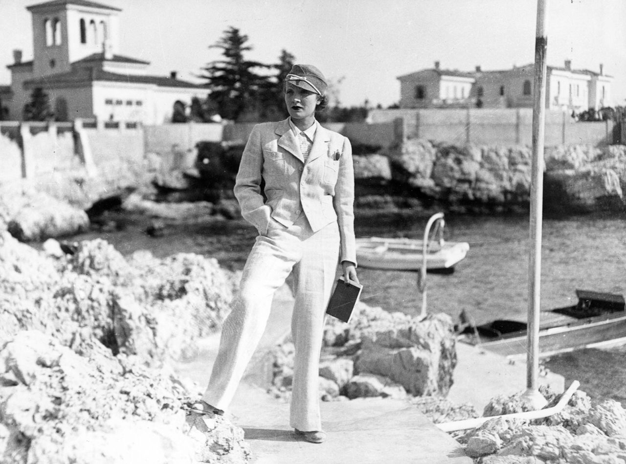 Actress and cabaret performer Marlene Dietrich poses in a circa-1932 photograph, standing on some coastal rocks. She is wearing a tailored suit and pillbox hat.