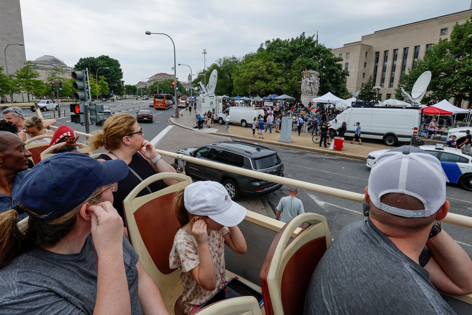 Tourists atop a double-decker tour bus pass by assembled reporters, law enforcement officers and demonstrators as they wait for Trump to arrive at the federal courthouse.