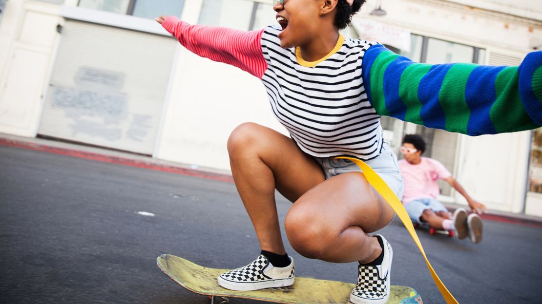 An excited young woman wearing a colorful outfit riding a skateboard down a city street