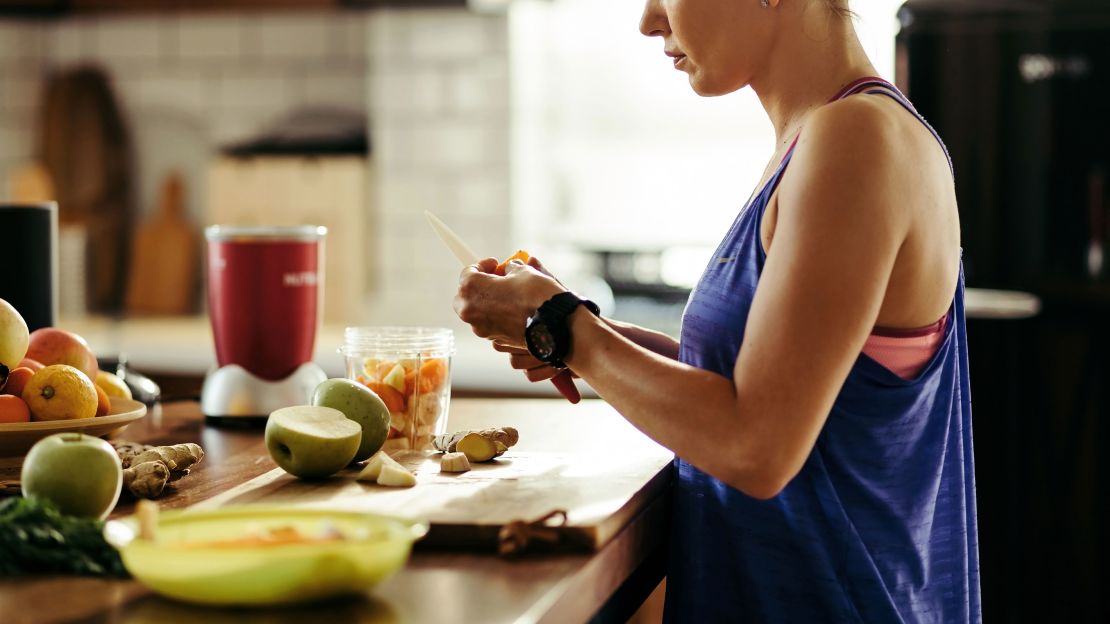 Young sportswoman making herself a healthy smoothie and slicing fresh fruit in the kitchen.