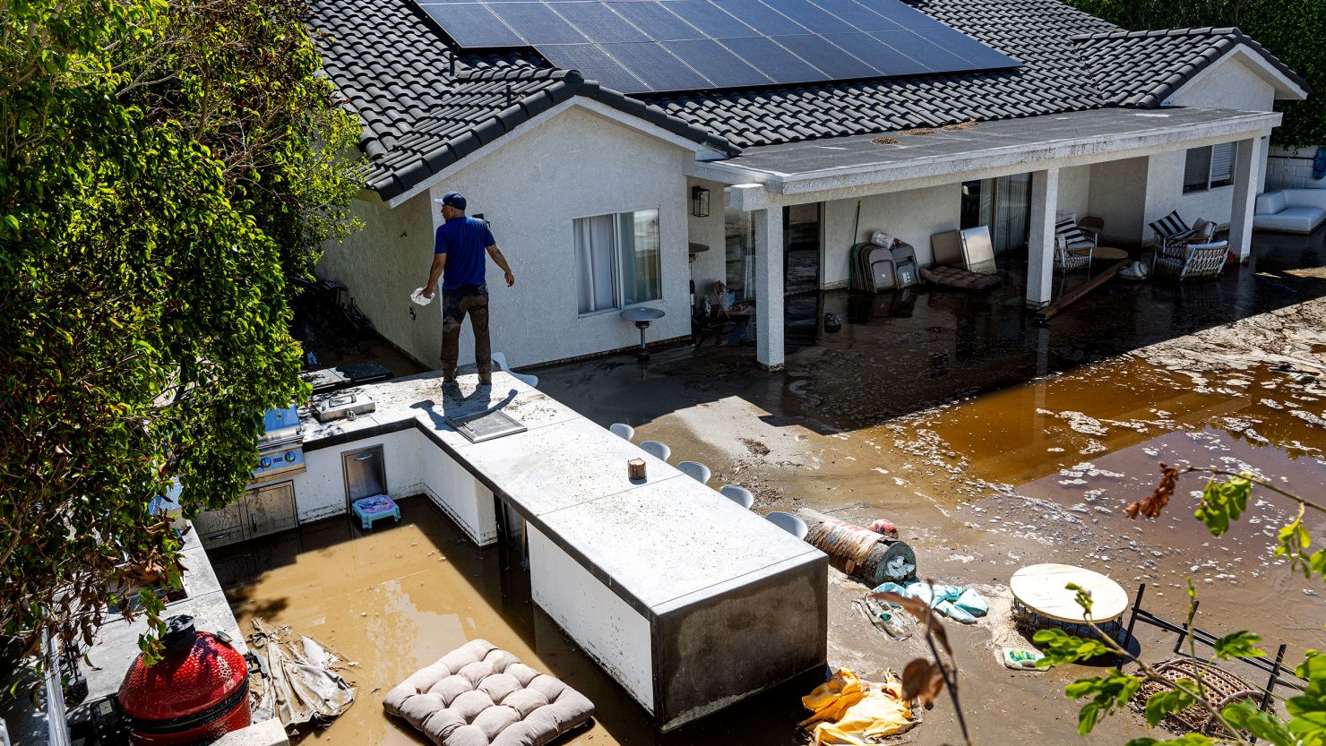 CATHEDRAL CITY, CA  - AUGUST 21, 2023: Nick Rodriguez stands on top of an outdoor barbecue seating area  at his brother's flooded backyard from the remnants of tropical storm Hilary on August 21, 2023 in Cathedral City, California. The inside of the house has 18 inches of flood water. "The water came into the house about midnight full throttle, Rick Figueroa said. He and his family hunkered down in one room until sunrise. Unable to escape out their front door Figueroa's wife Dawn and daughter Ella escaped over a neighbor's back wall Monday morning.(Gina Ferazzi / Los Angeles Times via Getty Images)