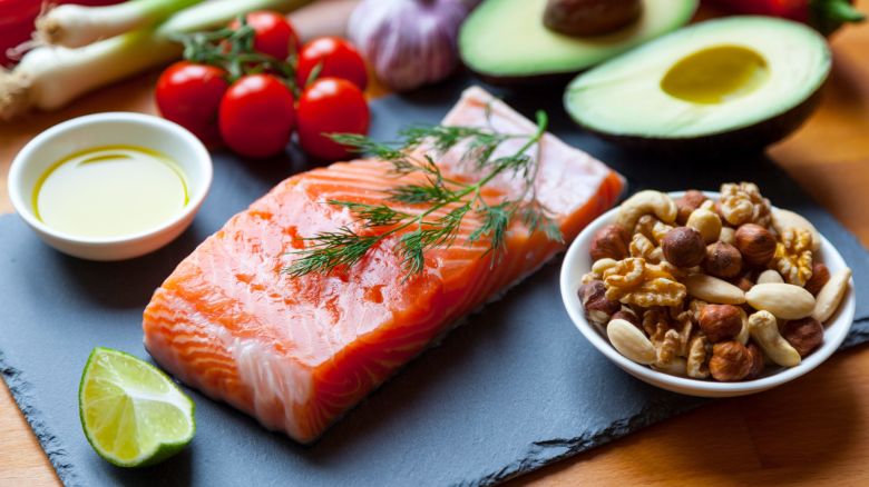 Table top still life of foods high in healthy fats such as Salmon, olive oil, nuts and avocados with vegetables and herbs.