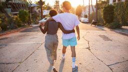 A hip young African American couple with their arms around each other walking down a suburban street at sunset shot from behind