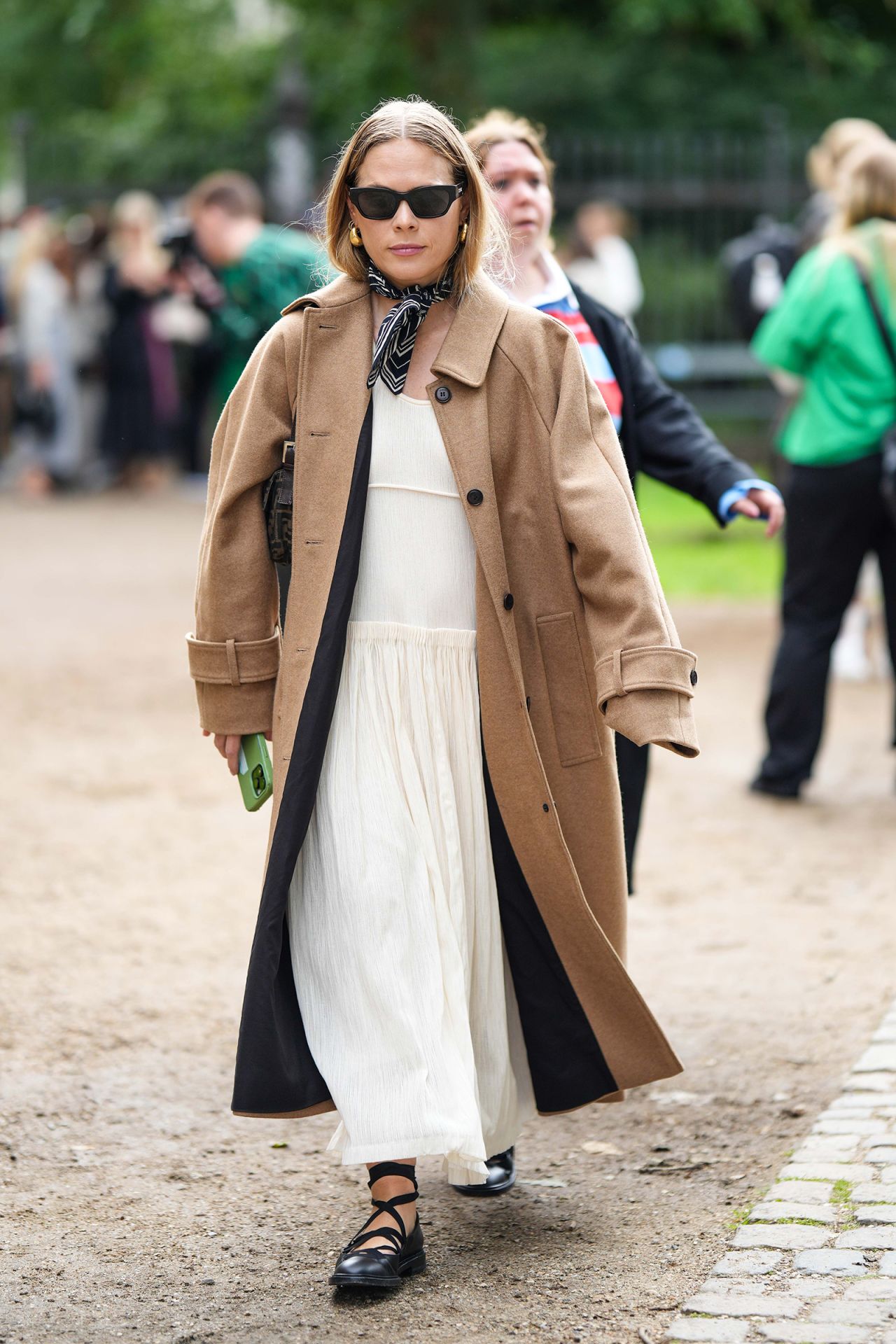 COPENHAGEN, DENMARK - AUGUST 10: A guest wears black sunglasses from Celine, gold earrings, a navy blue and white print pattern silk scarf, a beige brown long wool coat, a black long kimono, a white latte pleated / accordion long dress, black shiny leather laces ballerinas shoes , a brown and black FF monogram print pattern denim shoulder bag from Fendi , outside Mark Kenly Domino Tan, during the Copenhagen Fashion Week Spring/Summer 2024 on August 10, 2023 in Copenhagen, Denmark. (Photo by Edward Berthelot/Getty Images)