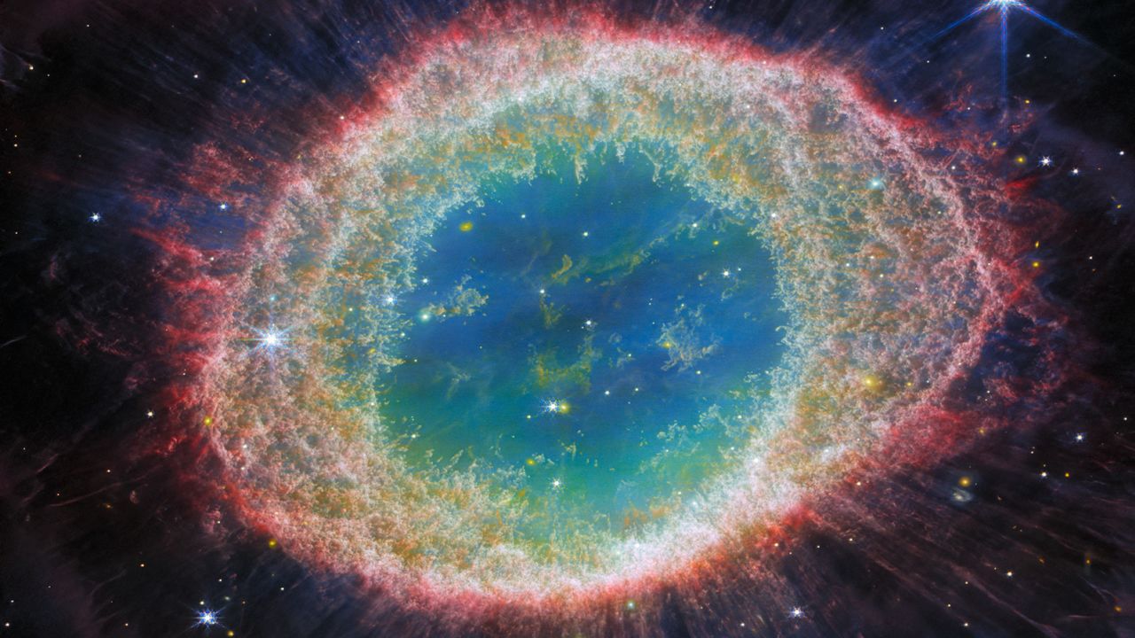 The NASA/ESA/CSA James Webb Space Telescope has observed the well-known Ring Nebula with unprecedented detail. Formed by a star throwing off its outer layers as it runs out of fuel, the Ring Nebula is an archetypal planetary nebula. Also known as M57 and NGC 6720, it is both relatively close to Earth at roughly 2,500 light-years away. This new image provides unprecedented spatial resolution and spectral sensitivity. For example, the intricate details of the filament structure of the inner ring are particularly visible in this dataset. There are some 20,000 dense globules in the nebula, which are rich in molecular hydrogen. In contrast, the inner region shows very hot gas. The main shell contains a thin ring of enhanced emission fromcarbon-based molecules known as polycyclic aromatic hydrocarbons (PAHs). Roughly ten concentric arcs are located just beyond the outer edge of the main ring. The arcs are thought to originate from the interaction of the central star with a low-mass companion orbiting at a distance comparable to that between the Earth and the dwarf planet Pluto. In this way, nebulae like the Ring Nebula reveal a kind of astronomical archaeology, as astronomers study the nebula to learn about the star that created it. [Image description: This image of the Ring Nebula appears as a distorted doughnut. The nebula's inner cavity hosts shades of blue and green, while the detailed ring transitions through shades of orange in the inner regions and pink in the outer region. The ring's inner region has distinct filament elements.]