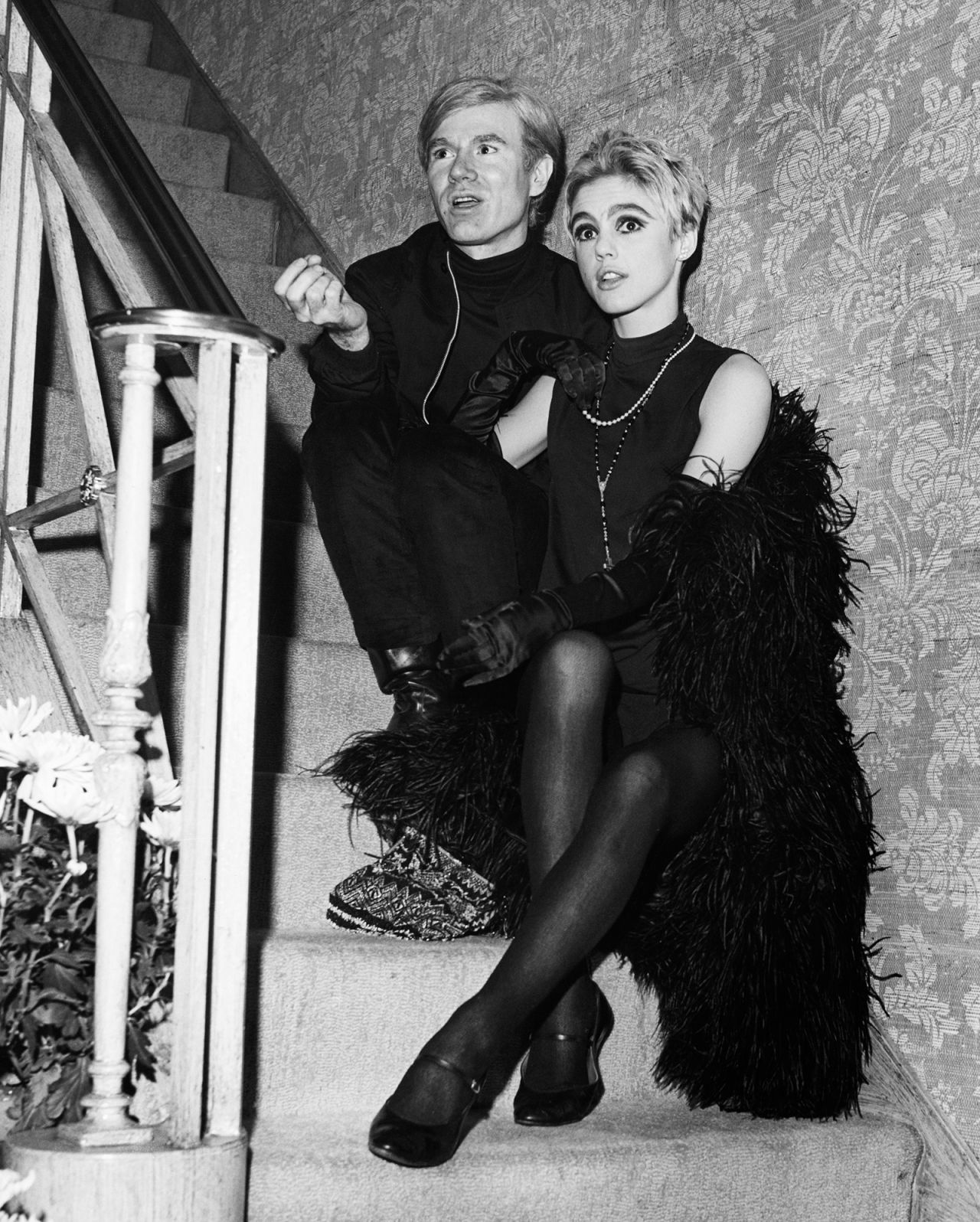 Художниците Анди Уорхол и Еди Седжуик, облечени в боа от черни пера, седят на стълбите и разговарят. 