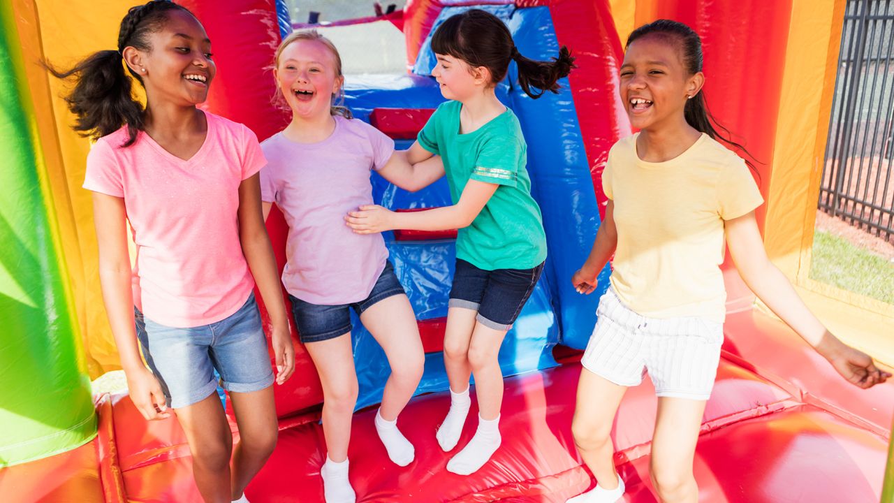 A multi-ethnic group of six girls playing together in a bounce house, smiling and laughing. The girl second from the left has down syndrome.