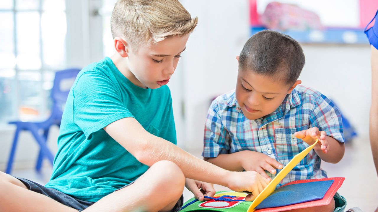 An elementary age Caucasian boy helps read a book to a preschool age Hispanic boy with Down Syndrome. They are looking down at the book and enjoying each other's company.