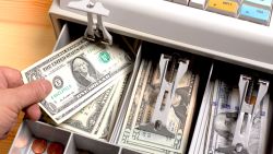 close up shot of the open drawer of a cash till, full of American dollars.  Sales assistant giving change to customer