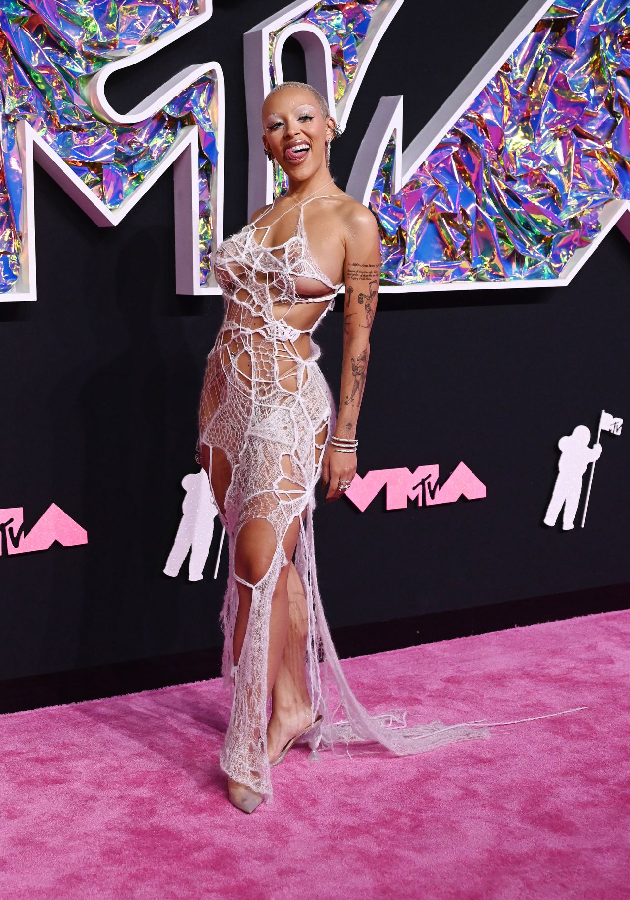 Doja Cat wore a daring Ap0cene gown paired with see-through pointed pumps, diamond chandelier earrings and glittery wraparound silver bracelets. Her makeup continued the theme with spidery black eyelashes and a bold white eyeshadow and thin arched brows. 