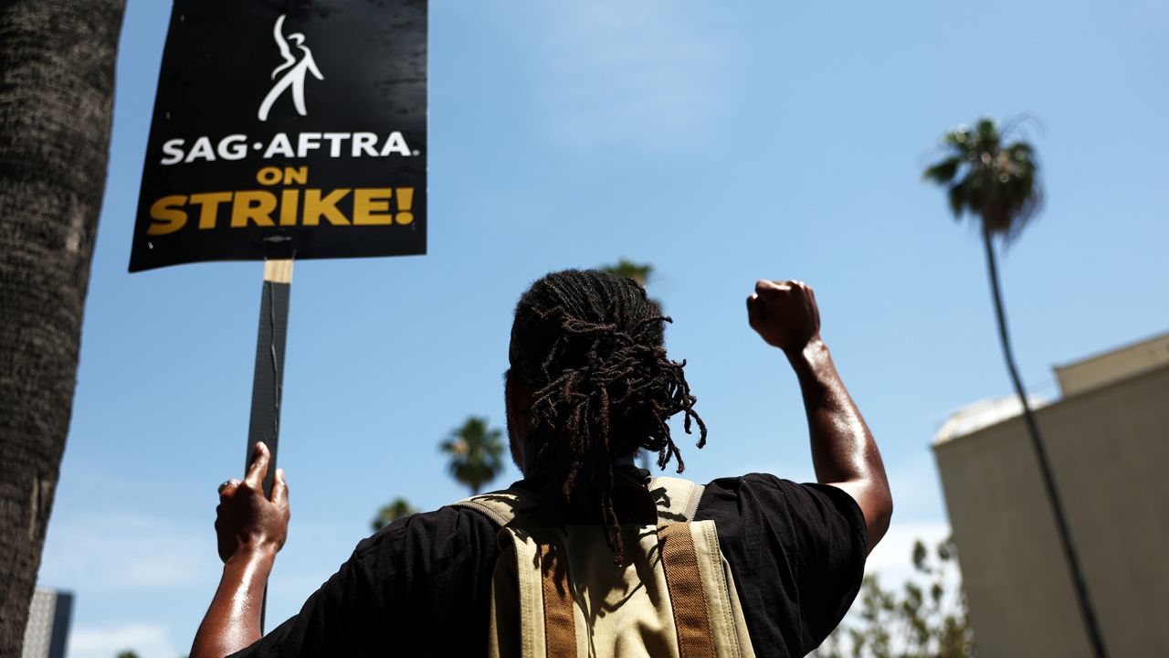 एक हड़ताली SAG-AFTRA सदस्य अन्य SAG-AFTRA सदस्यों और हड़ताली WGA (राइटर्स गिल्ड ऑफ अमेरिका) के कार्यकर्ताओं के साथ 17 जुलाई को कैलिफोर्निया के बरबैंक में वार्नर ब्रदर्स स्टूडियो के बाहर धरना देता है। 