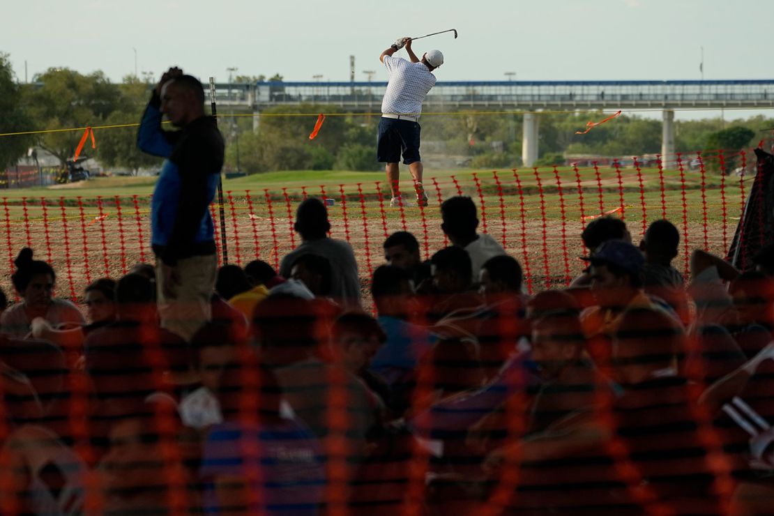 Migrants who crossed into the US from Mexico watch a golfer from a makeshift processing area along the Rio Grande, on Thursday, Sept. 21, 2023, in Eagle Pass, Texas.