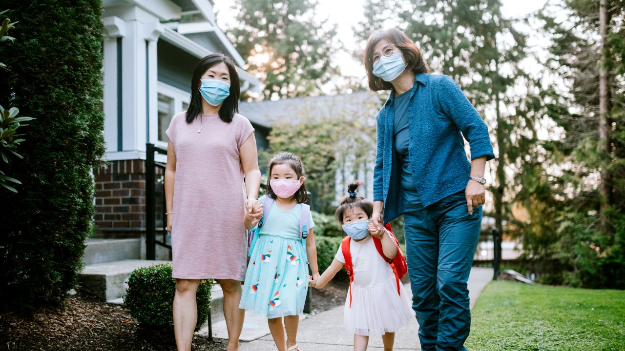 A Korean woman helps her two small daughters get ready to go to school, their grandmother assisting as well.  The girls wearing protective face masks and backpacks.  Adjusting to the new normal of Covid-19 / Coronavirus