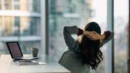 Back view of businesswoman looking through the window while relaxing with hands behind head in the office. Copy space.