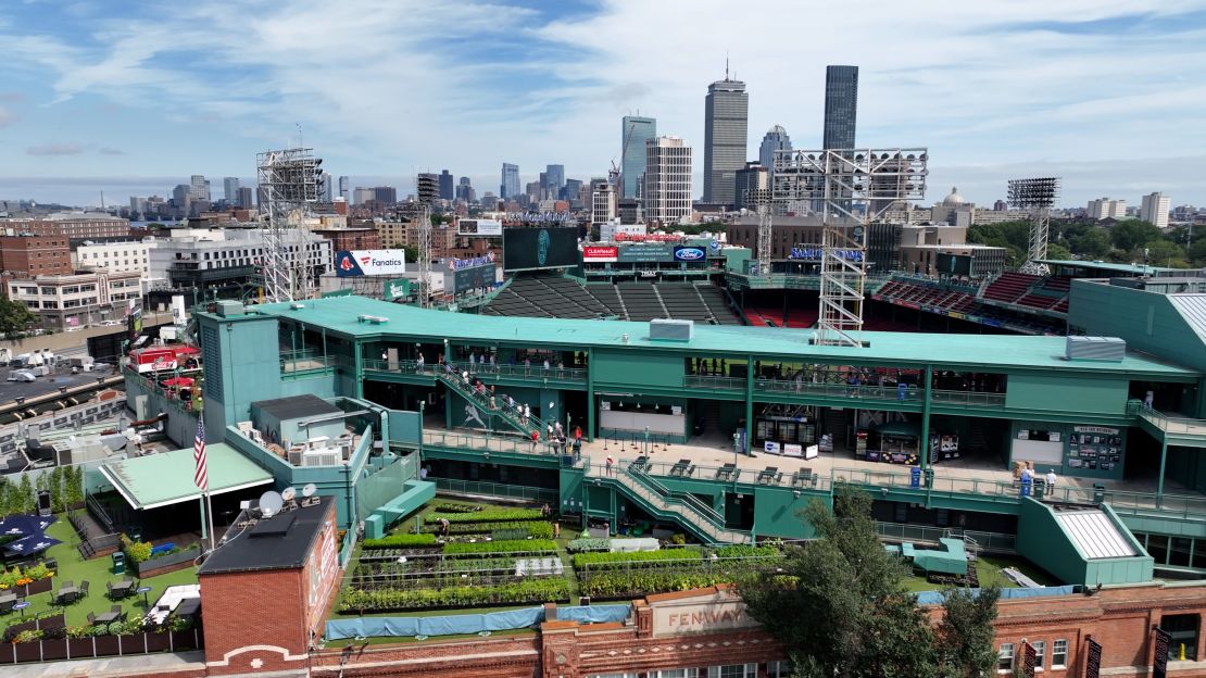 An aerial view of Fenway Park shows Fenway Farms in the foreground, against a backdrop of the Boston skyline.