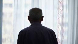 Silhouette of retired man looking through window with transparent curtain standing at home rear view. Loneliness and old human care concept.