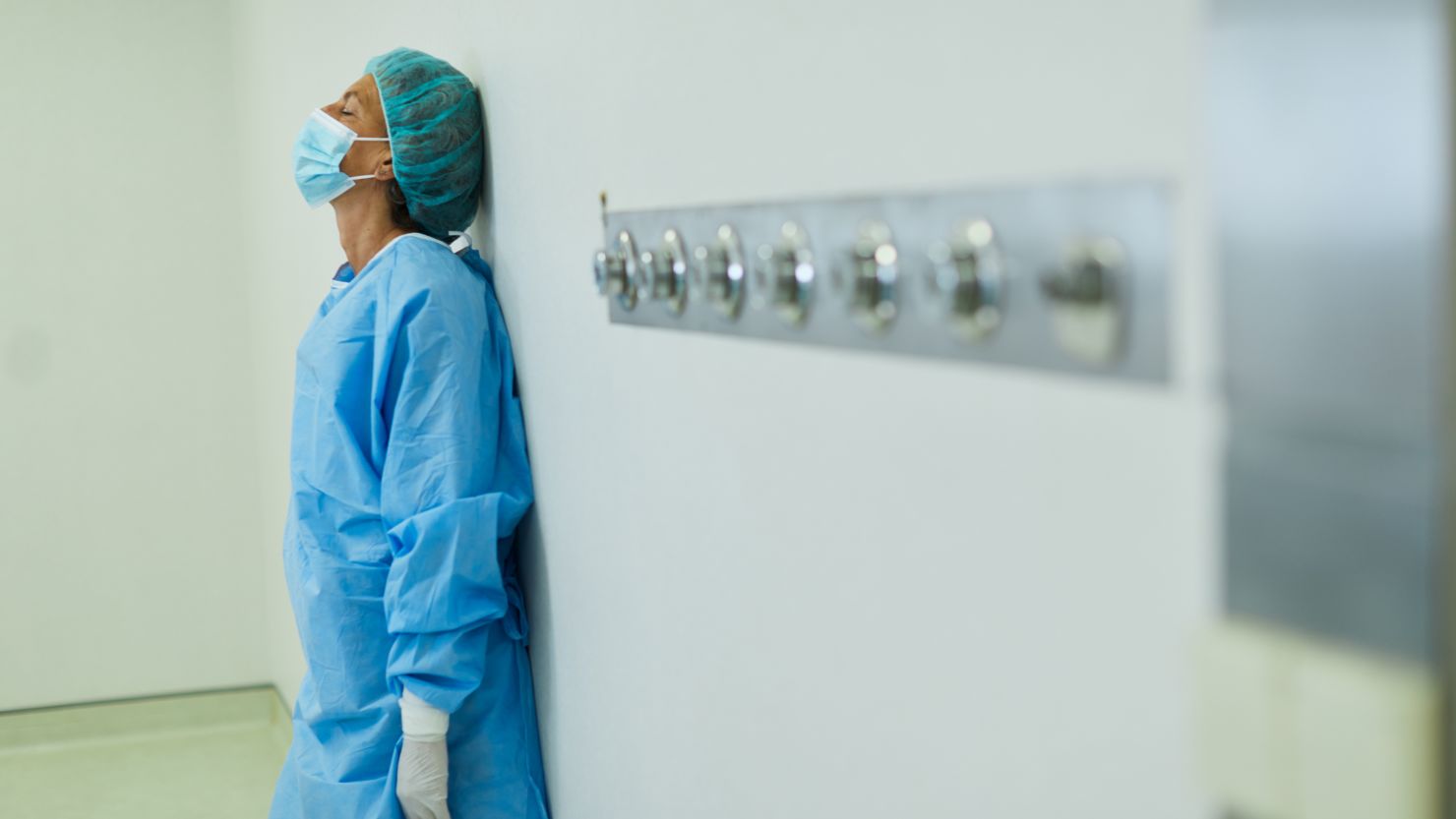 Saddened by an  unsuccessful operation this tired surgical nurse still wearing her surgical gown, mask, cap and gloves takes a moment to recover by leaning back against the wall for support. Blurred right side, three quarter length with copy space.