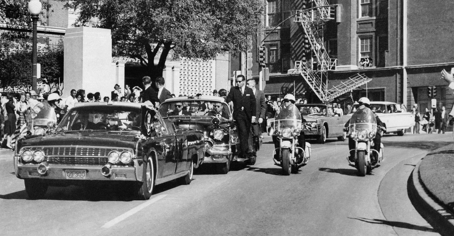 The motorcade proceeds along Elm Street past the Texas School Book Depository, shortly after Kennedy was shot. Looking through the limo's windshield in the foreground, Kennedy appears to raise his hand toward his head after being shot. The first lady holds his forearm in an effort to aid him.