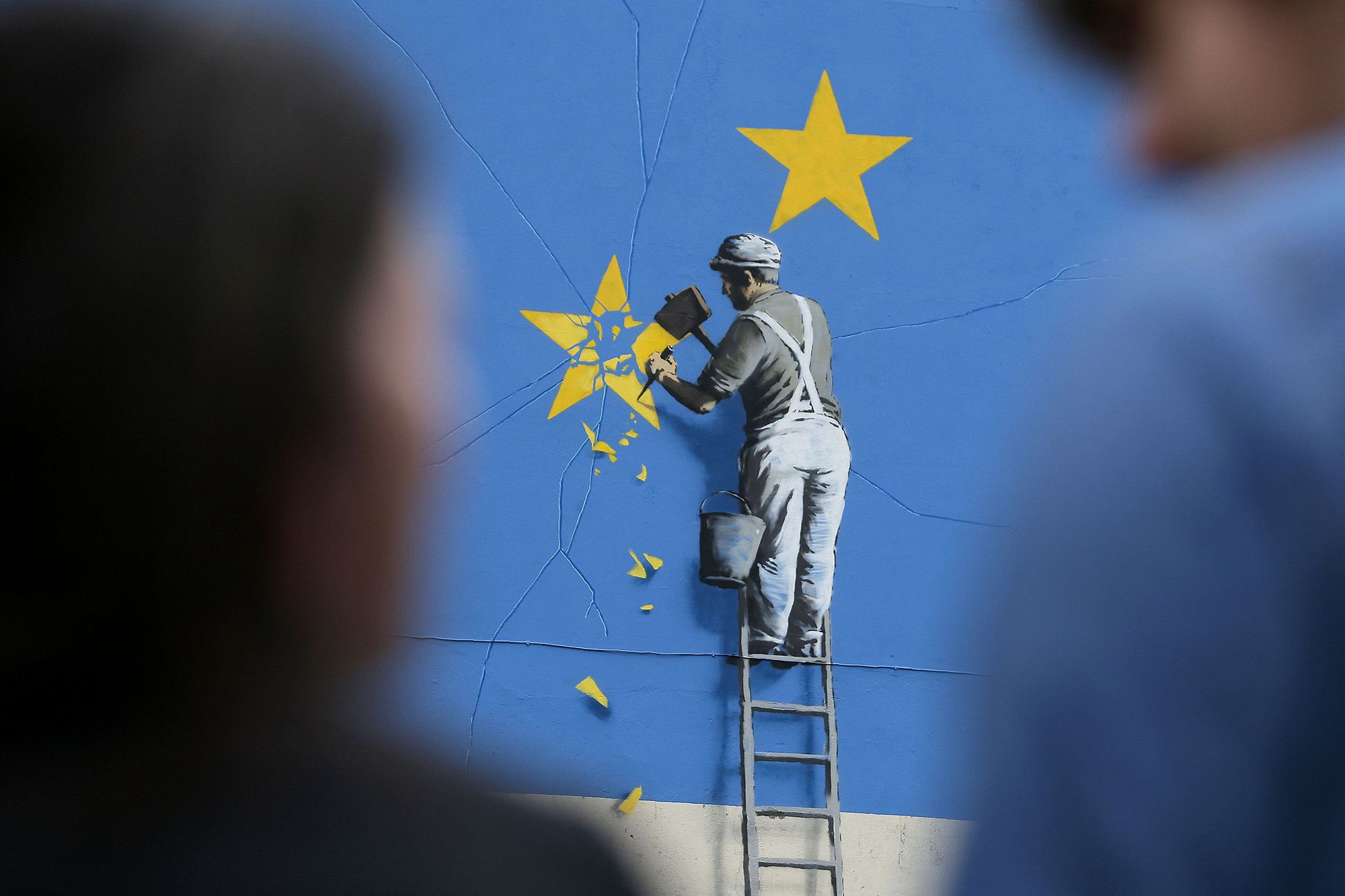 People stand near a recently painted mural by British graffiti artist Banksy, depicting a workman chipping away at one of the stars on a European Union (EU) themed flag, in Dover, south east England on May 8, 2017. (Photo by Daniel LEAL / AFP) / RESTRICTED TO EDITORIAL USE - MANDATORY MENTION OF THE ARTIST UPON PUBLICATION - TO ILLUSTRATE THE EVENT AS SPECIFIED IN THE CAPTION (Photo by DANIEL LEAL/AFP via Getty Images)