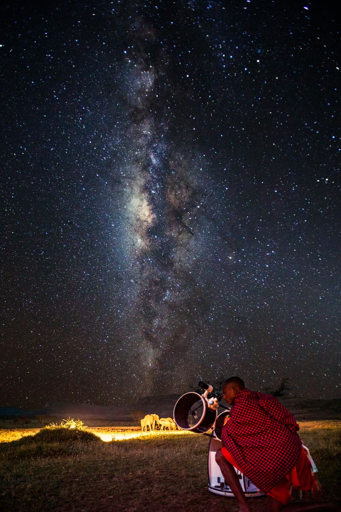 A Masaai man watches the stars, while nearby elephants look on.