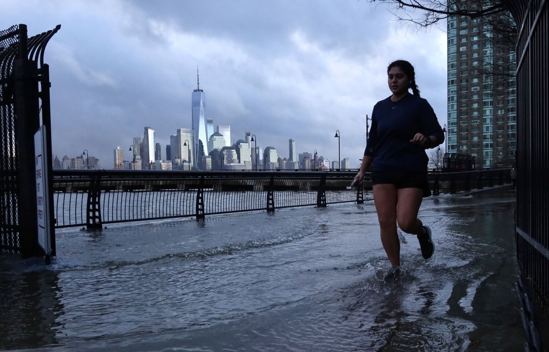 Water from the Hudson River overflows a river wall at high tide as a woman jogs in front of the skyline of lower Manhattan in New York City on January 13, 2024, in Jersey City, New Jersey.