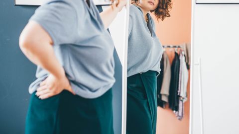 Young woman in glasses admires choice of clothes standing in front of large mirror in stylish room reflection view.