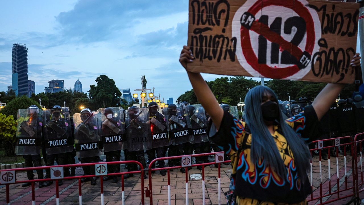 A protester holds a sign referencing article 112, Thailand's lese majeste royal defamation law, as riot police keep watch during a demonstration in Bangkok on November 14, 2021, after a Thai court ruled that speeches by protest leaders calling for royal reforms amounted to a bid to overthrow the country's monarchy.