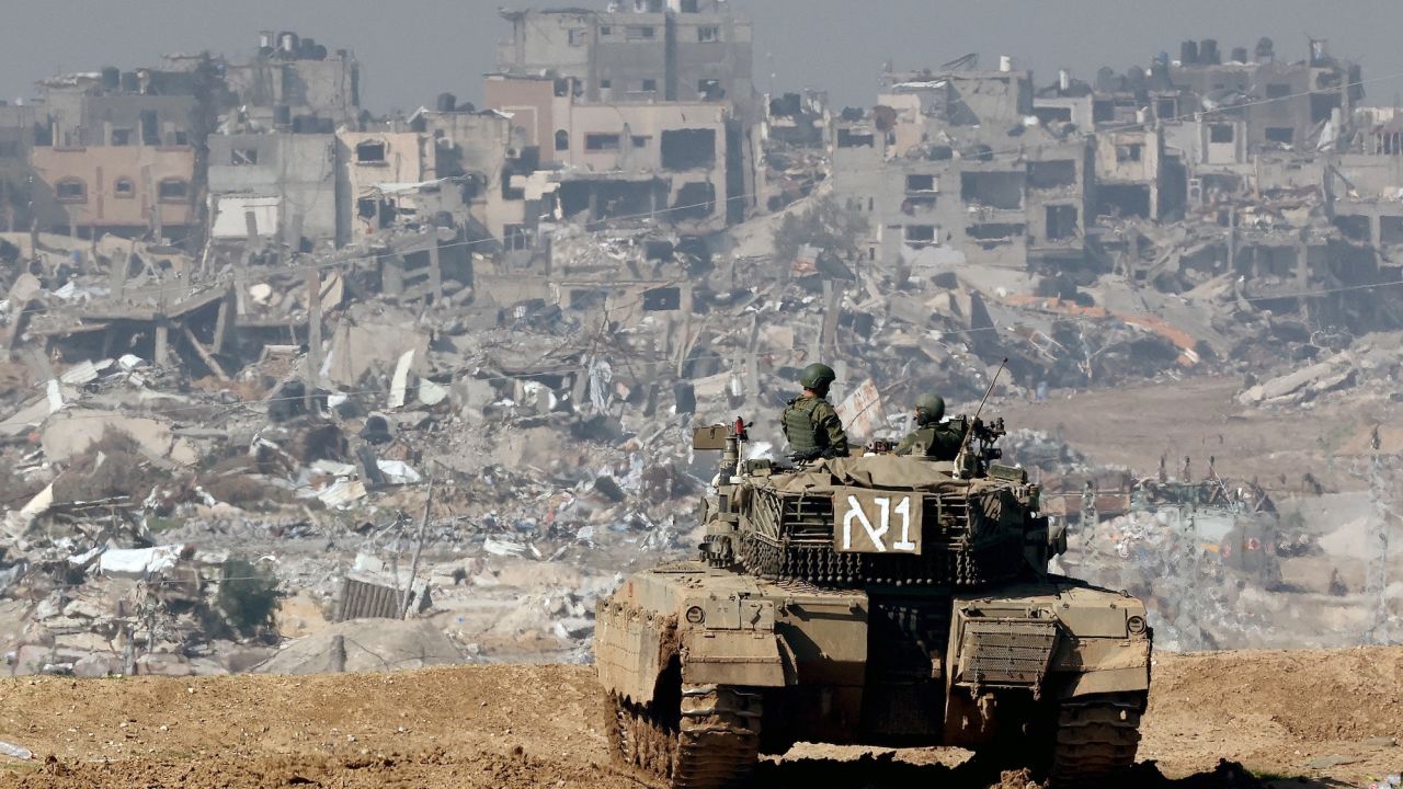 A picture taken from a position in southern Israel along the border with the Gaza Strip on January 19, 2024, shows an Israeli tank rolling along the fence as damaged buildings are see in the Gaza strip amid continuing battles between Israel and the militant group Hamas. (Photo by JACK GUEZ / AFP)