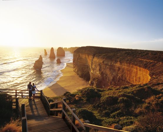 Man and woman admiring the sea at sunset.  In the distance are the Twelve Apostles, unique rock formations that rise out of the sea along the Victorian coast.