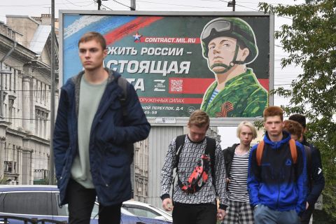 Los hombres jóvenes caminan frente a una valla publicitaria que promueve el servicio militar por contrato con la imagen de un militar y el eslogan que dice 
