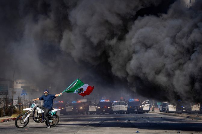 A man on a motorcycle waves a Mexican flag as smoke rises from a burning car during <a  target="_top" href="/newspapers?url=https://www.cnn.com/2025/06/09/us/gallery/los-angeles-protests">protests in Los Angeles</a> on Saturday, June 7.