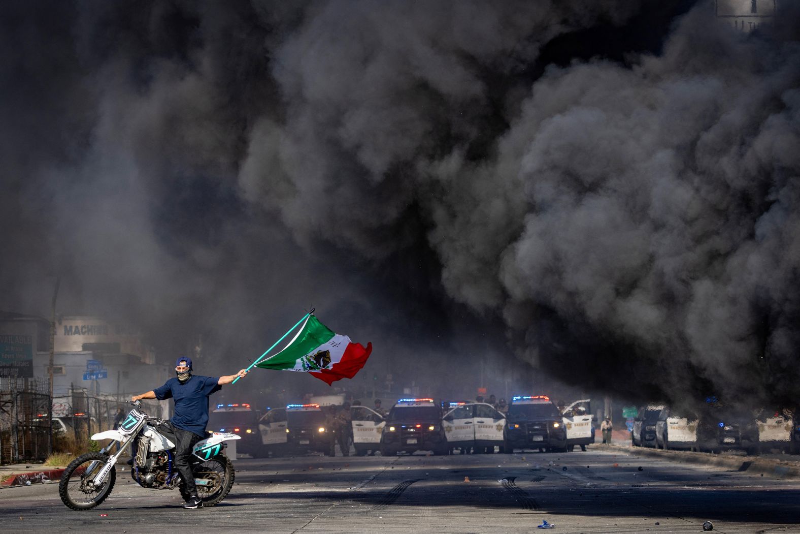 A man on a motorcycle waves a Mexican flag as smoke rises from a burning car during <a href="index.php?page=&url=https%3A%2F%2Fwww.cnn.com%2F2025%2F06%2F09%2Fus%2Fgallery%2Flos-angeles-protests">protests in Los Angeles</a> on Saturday, June 7.