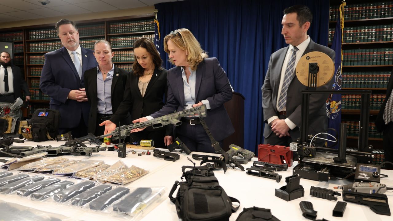Queens District Attorney Melinda Katz (center, holding gun) stands behind a display of the weapons and supplies uncovered in the raid.