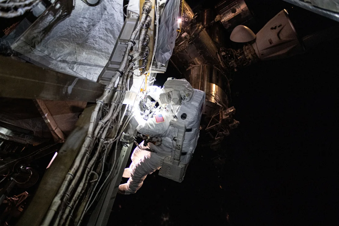 Un astronauta con un traje espacial blanco y la bandera de EE.UU. en su brazo realiza una caminata espacial mientras trabaja en la estructura de la Estación Espacial Internacional (ISS). En el fondo, se pueden ver módulos y componentes de la estación flotando en el espacio.