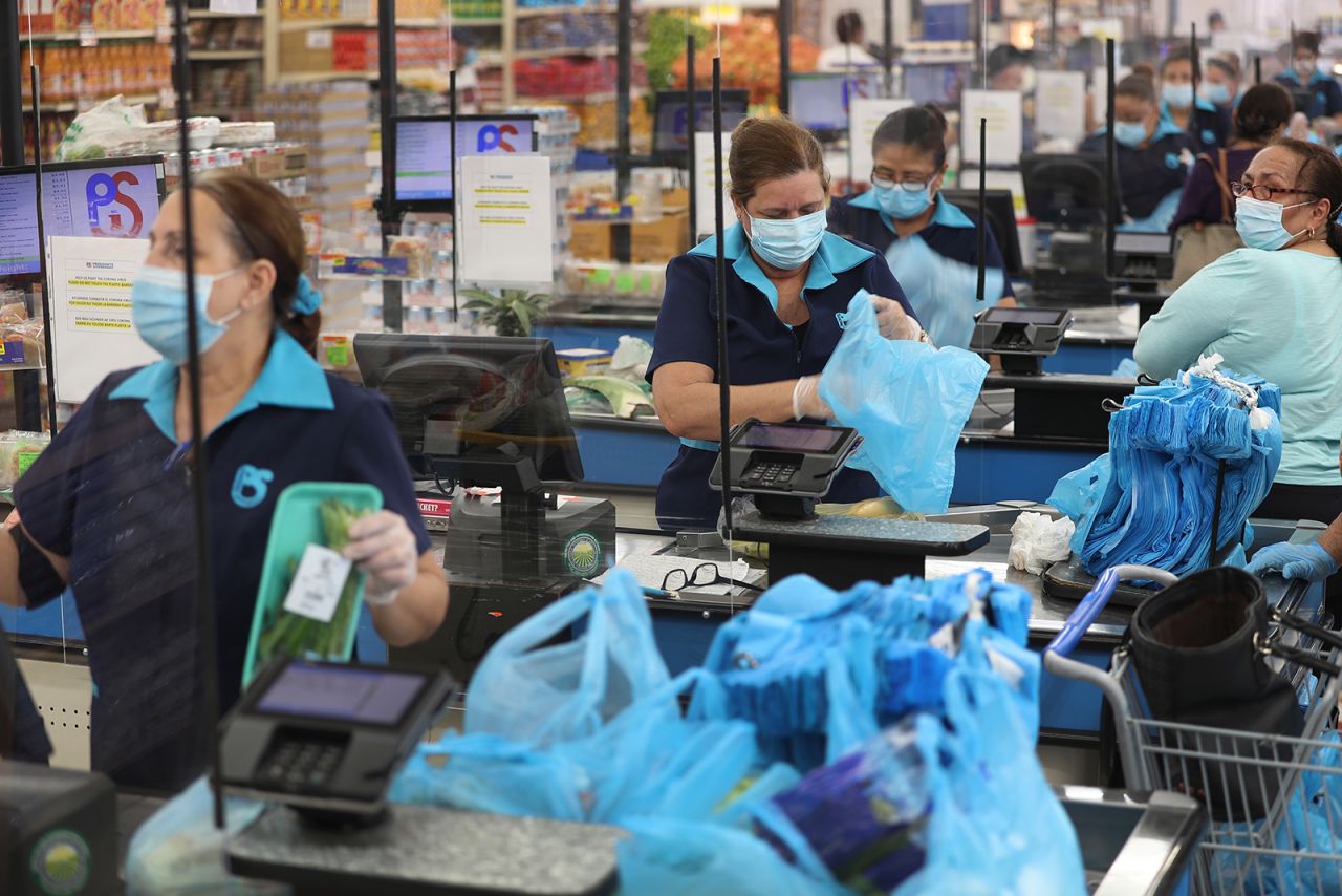 Cashiers at a grocery store in stand behind a partial protective plastic screen and wear masks and gloves as they ring up customers in Miami, Florida, on April 13.