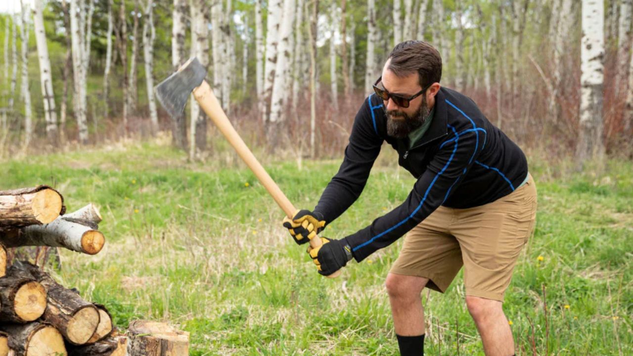 A bearded person with sunglasses black quarter-zip chops wood in a grass clearing. A pile of logs is on the left.