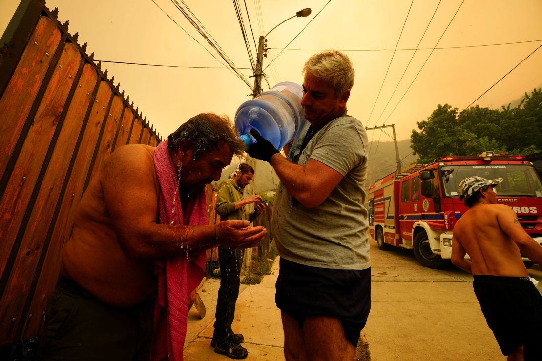 A man helps a fellow resident cool down with bottled water as forest fires burn nearby.