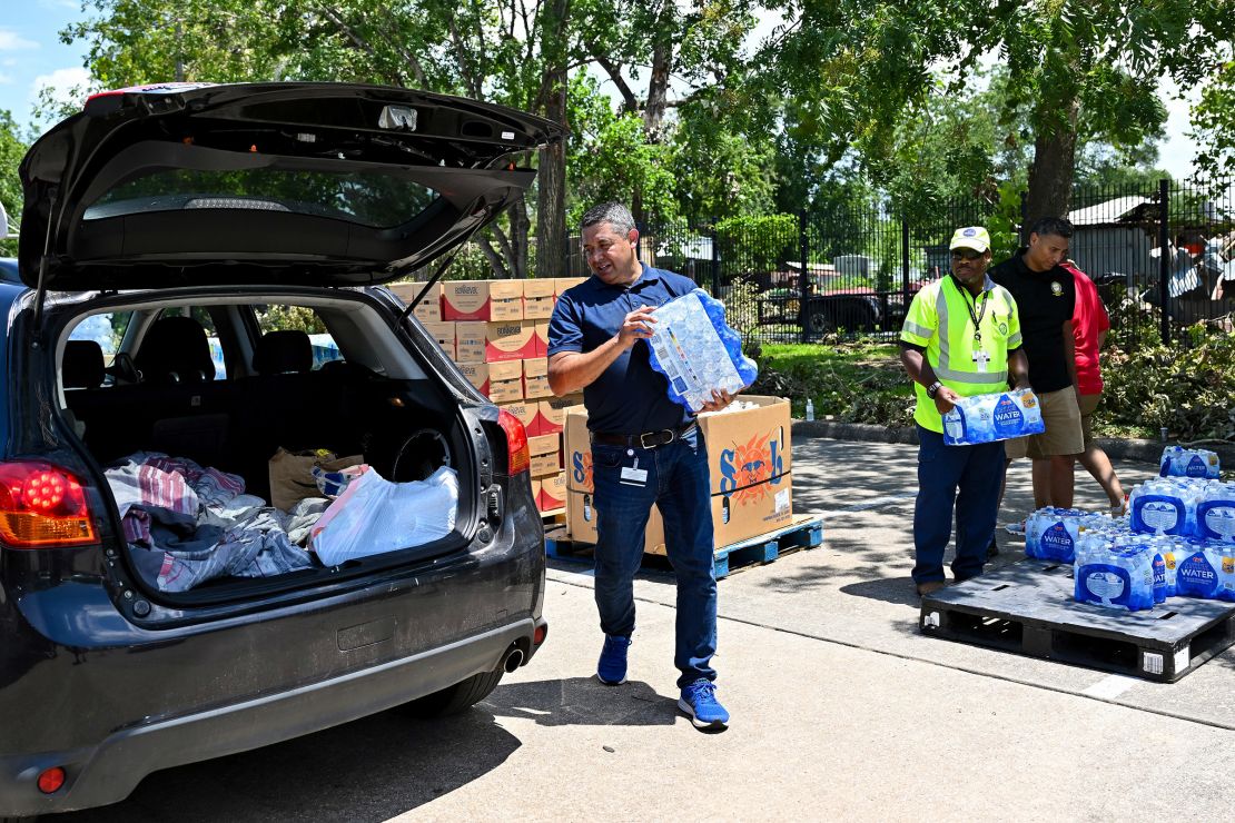 A man carries a case of water bottles toward the open trunk of a van.